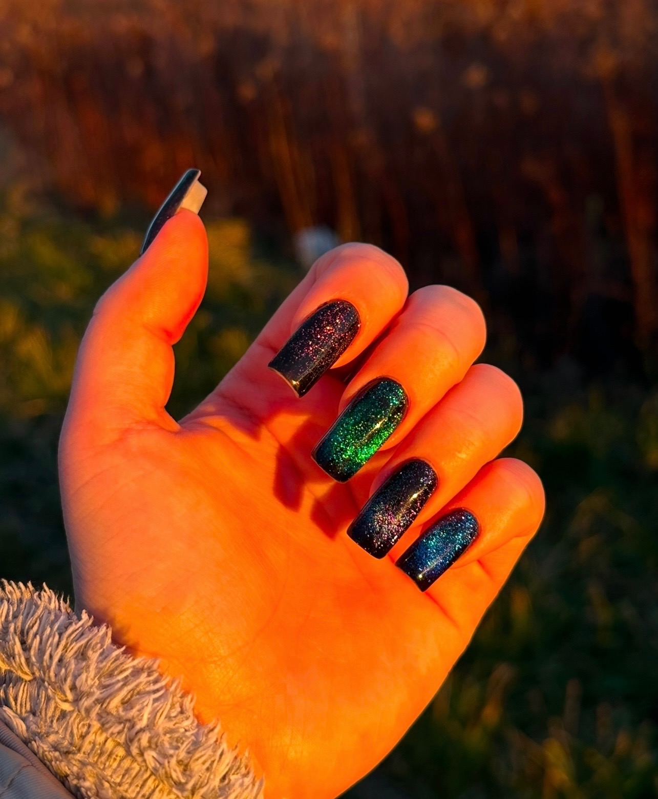 Dłoń z długimi, kwadratowymi paznokciami w odcieniach czerni, zieleni i błękitu z brokatowym wykończeniem, oświetlona ciepłym światłem zachodzącego słońca.