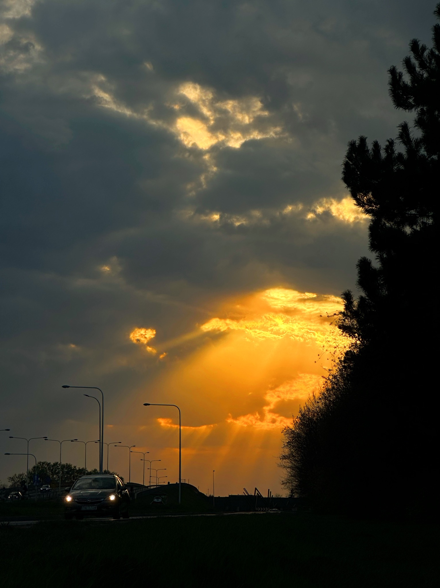 Pejzaż miejski o zmierzchu: promienie słońca przebijają się przez ciemne chmury, oświetlając drogę z jadącym samochodem i rzędem latarni. Silueta drzewa po prawej.