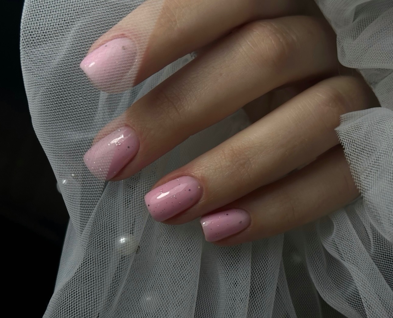 Delikatny manicure hybrydowy w odcieniach pudrowego różu z subtelnym brokatem, na tle białej, zwiewnej tkaniny. Krótkie, kwadratowe paznokcie.