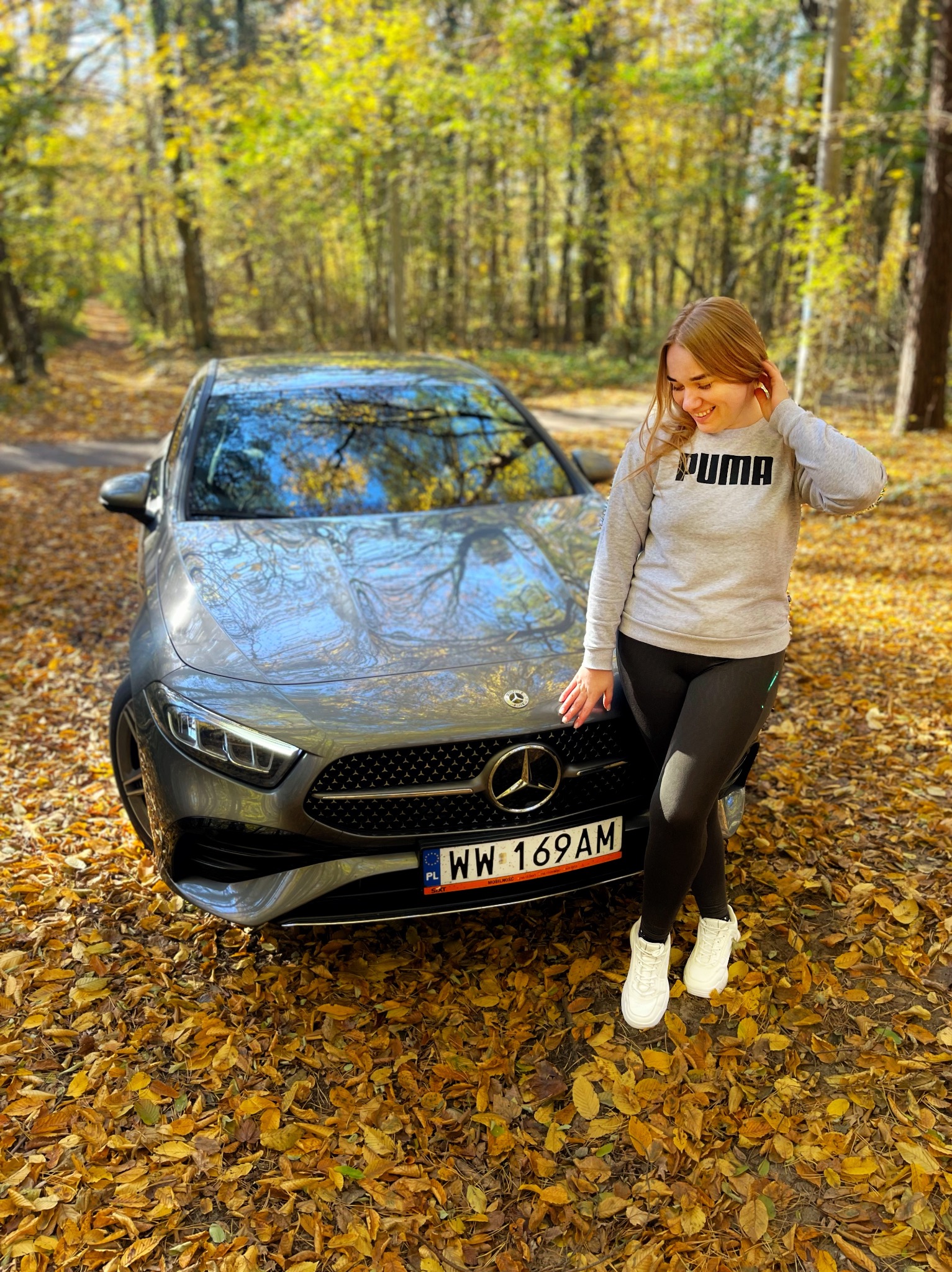Uśmiechnięta blondynka opiera się o szary samochód Mercedes na tle jesiennego lasu. Ziemię pokrywają opadłe, żółte liście. Widoczna tablica rejestracyjna.