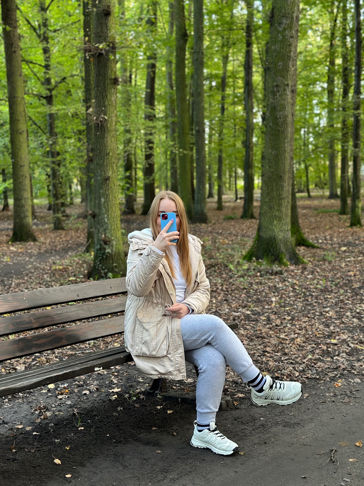 Kobieta w parku siedzi na ławce, trzymając niebieski smartfon przed twarzą. W tle drzewa i opadłe liście. Ubrana w beżową kurtkę i szare spodnie dresowe.