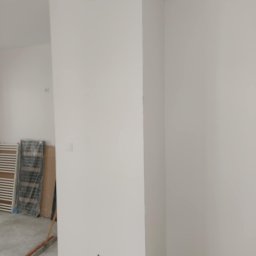 PI Development - Świeżo pomalowane na biało ściany w rogu pomieszczenia. Widoczny fragment podłogi, grzejnik w folii i szczotka. Minimalistyczne wykończenie wnętrza po malowaniu.