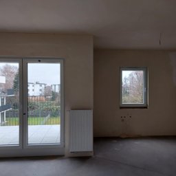 PI Development - Surowe wnętrze mieszkania po szpachlowaniu, z widokiem na balkon i grzejnikiem. Ściany przygotowane do malowania, surowa podłoga. Widok z okna na zewnątrz.