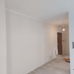 PI Development - Jasne, świeżo pomalowane wnętrze z widocznymi gniazdkami i listwą sufitową. W tle drzwi i krzesło, obok którego leżą narzędzia malarskie.