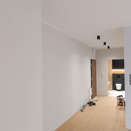 PI Development - Jasne, świeżo pomalowane wnętrze mieszkania z parkietem i widokiem na łazienkę. Minimalistyczne oświetlenie sufitowe i białe ściany tworzą nowoczesny wygląd.