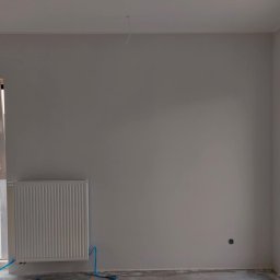 PI Development - Wnętrze pokoju po szpachlowaniu ścian na gładko, z grzejnikiem i oknem. Widoczne ślady prac wykończeniowych na podłodze i wystający kabel ze ściany.