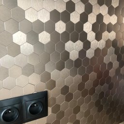 PI Development - Ściana wykończona metalicznymi, heksagonalnymi płytkami w odcieniach brązu i srebra. Widoczne gniazdka elektryczne w czarnej ramce. Nowoczesny design.