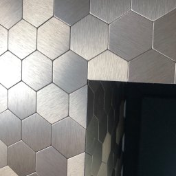 PI Development - Metaliczne heksagonalne płytki ścienne w odcieniach szarości, ułożone na ścianie, z fragmentem czarnej, błyszczącej powierzchni po prawej stronie.