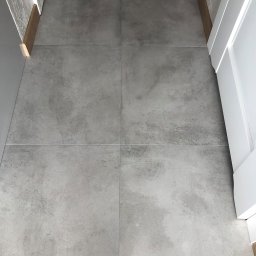 PI Development - Szare, matowe płytki podłogowe ułożone w kwadraty, widoczne białe fugi, przy ścianie drewniana listwa przypodłogowa, fragment białych drzwi po prawej.