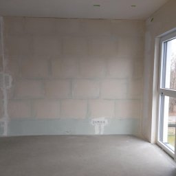 PI Development - Pokój po szpachlowaniu i gruntowaniu, przygotowany do malowania. Widoczne ślady po łatach gipsowych, gładzie i gniazdka elektryczne. Okno z widokiem na zewnątrz.