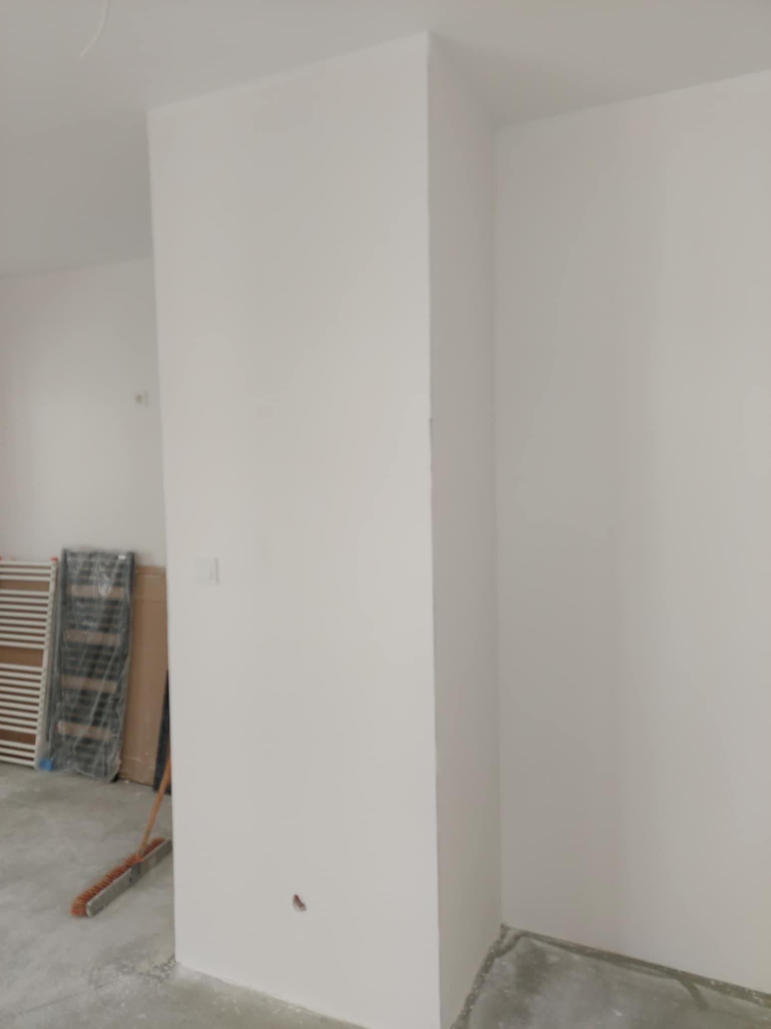 Świeżo pomalowane na biało ściany w rogu pomieszczenia. Widoczny fragment podłogi, grzejnik w folii i szczotka. Minimalistyczne wykończenie wnętrza po malowaniu.