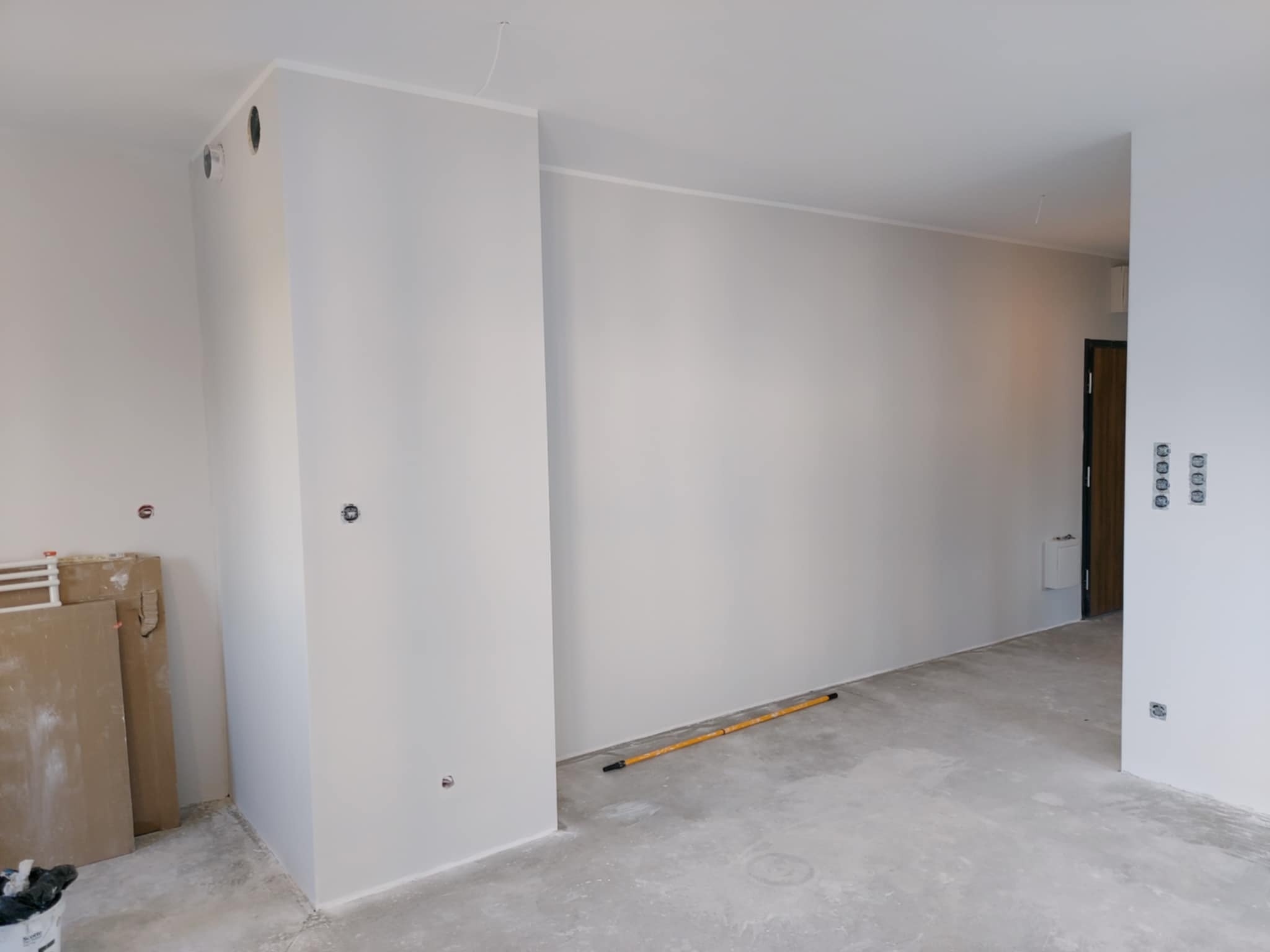 Wnętrze po malowaniu na szaro, widoczne gładkie ściany, surowa podłoga, narzędzie malarskie i płyty. Przygotowanie do dalszych prac wykończeniowych.