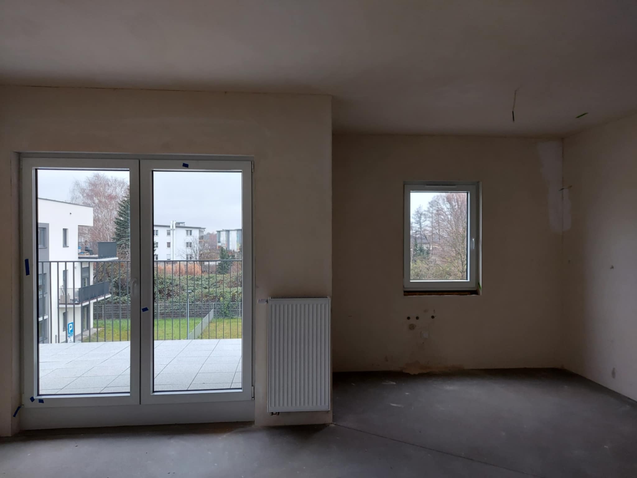 Surowe wnętrze mieszkania po szpachlowaniu, z widokiem na balkon i grzejnikiem. Ściany przygotowane do malowania, surowa podłoga. Widok z okna na zewnątrz.