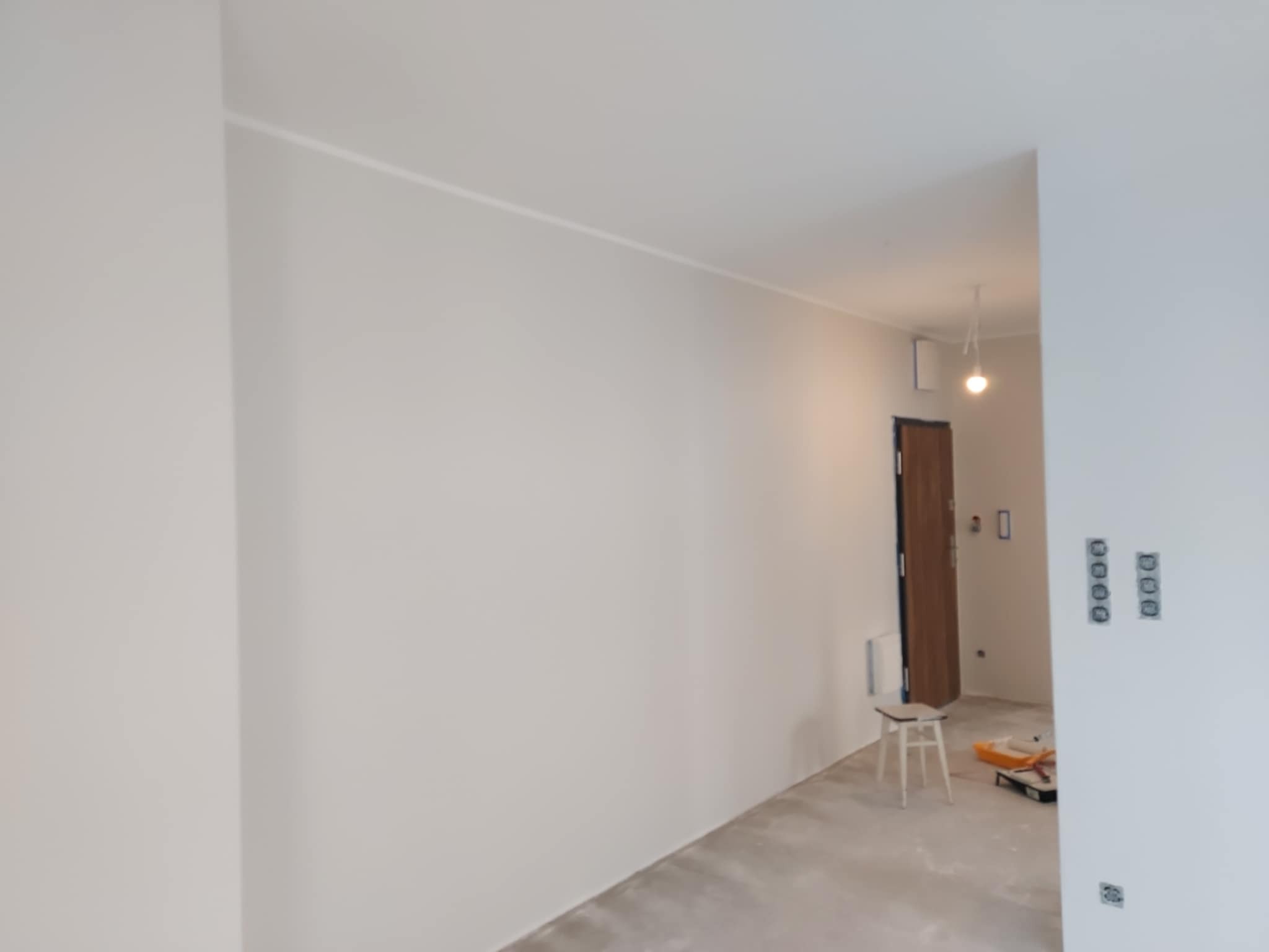 Jasne, świeżo pomalowane wnętrze z widocznymi gniazdkami i listwą sufitową. W tle drzwi i krzesło, obok którego leżą narzędzia malarskie.