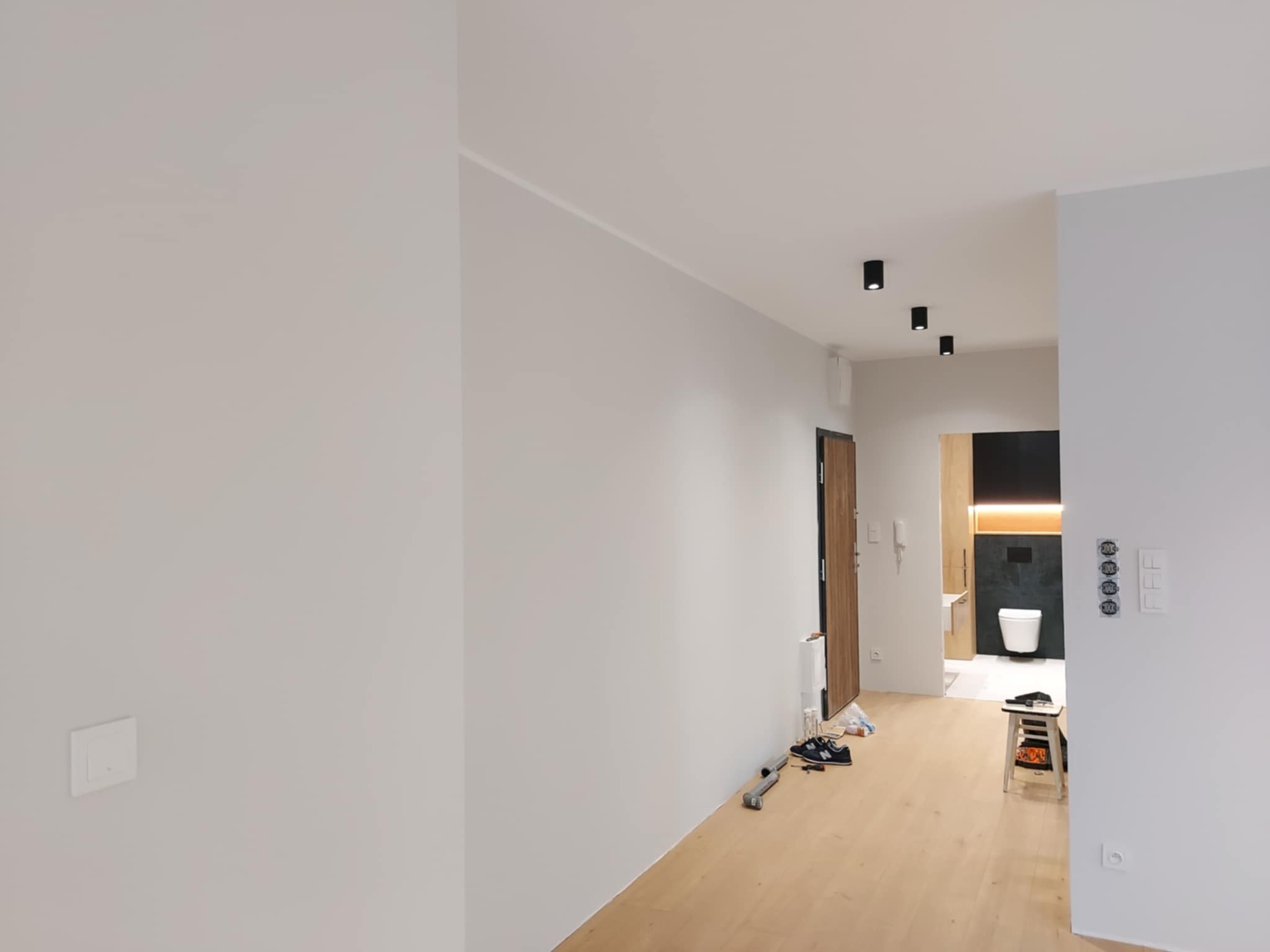 Jasne, świeżo pomalowane wnętrze mieszkania z parkietem i widokiem na łazienkę. Minimalistyczne oświetlenie sufitowe i białe ściany tworzą nowoczesny wygląd.