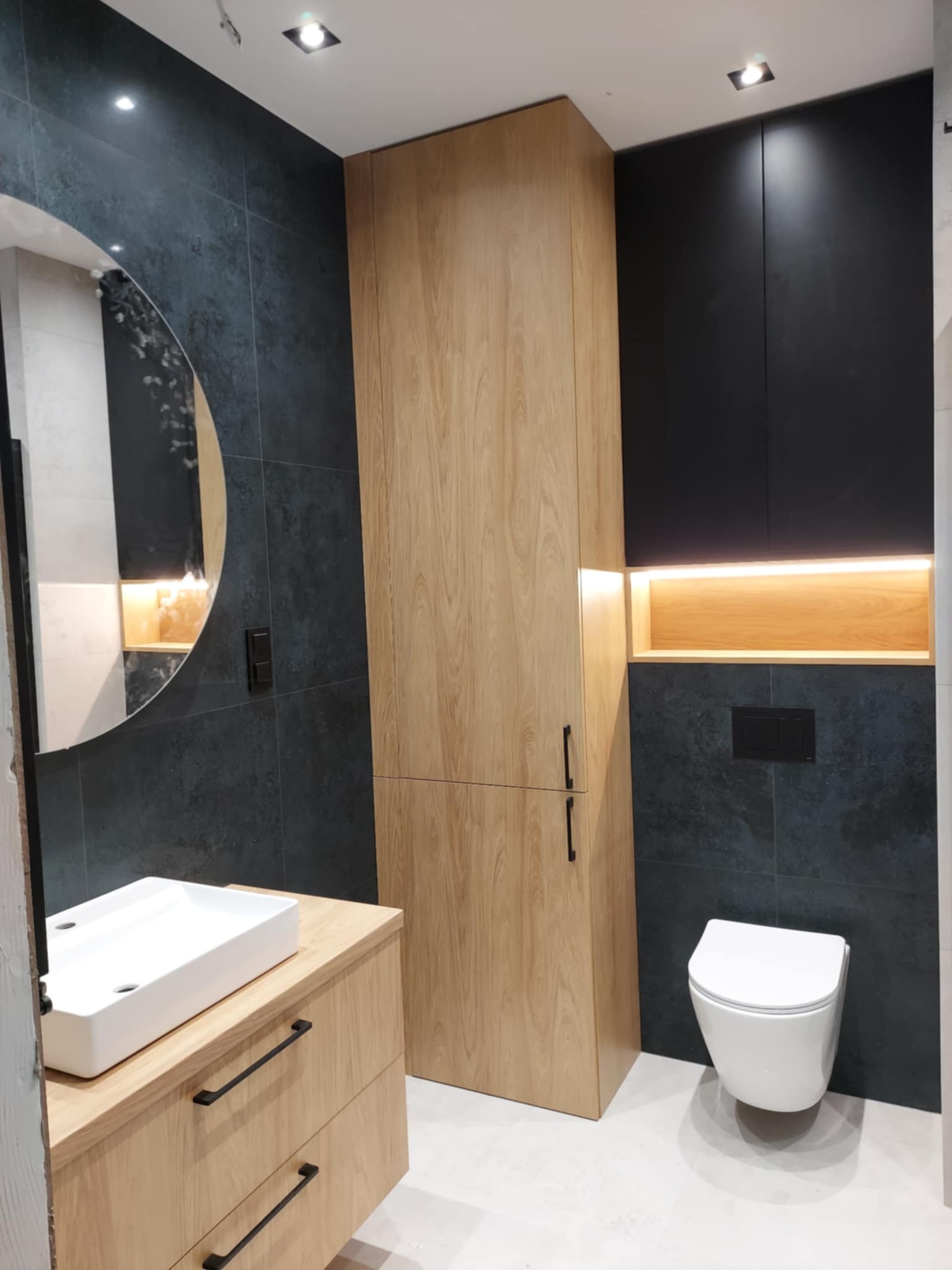 Nowoczesna łazienka z drewnianą szafką pod umywalką, okrągłym lustrem i podświetlaną wnęką nad wiszącą toaletą. Ciemne płytki na ścianach.