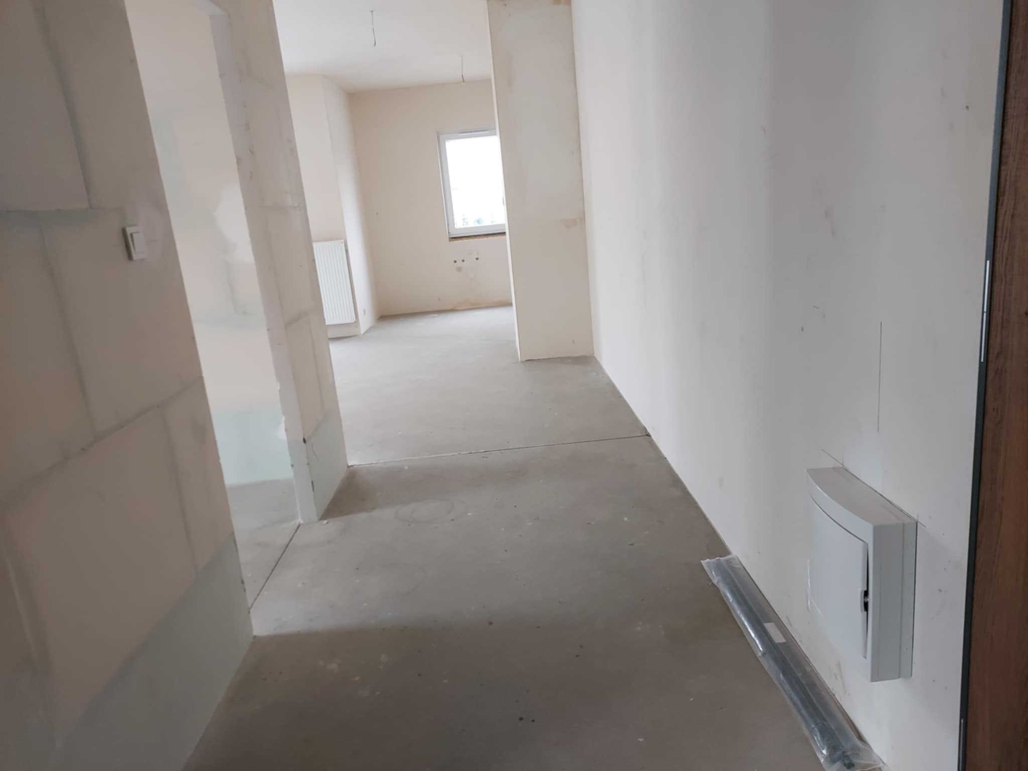 Surowe wnętrze w trakcie prac wykończeniowych. Widoczne ściany przygotowane do malowania, surowa podłoga i skrzynka elektryczna na ścianie. Jasne, naturalne światło.