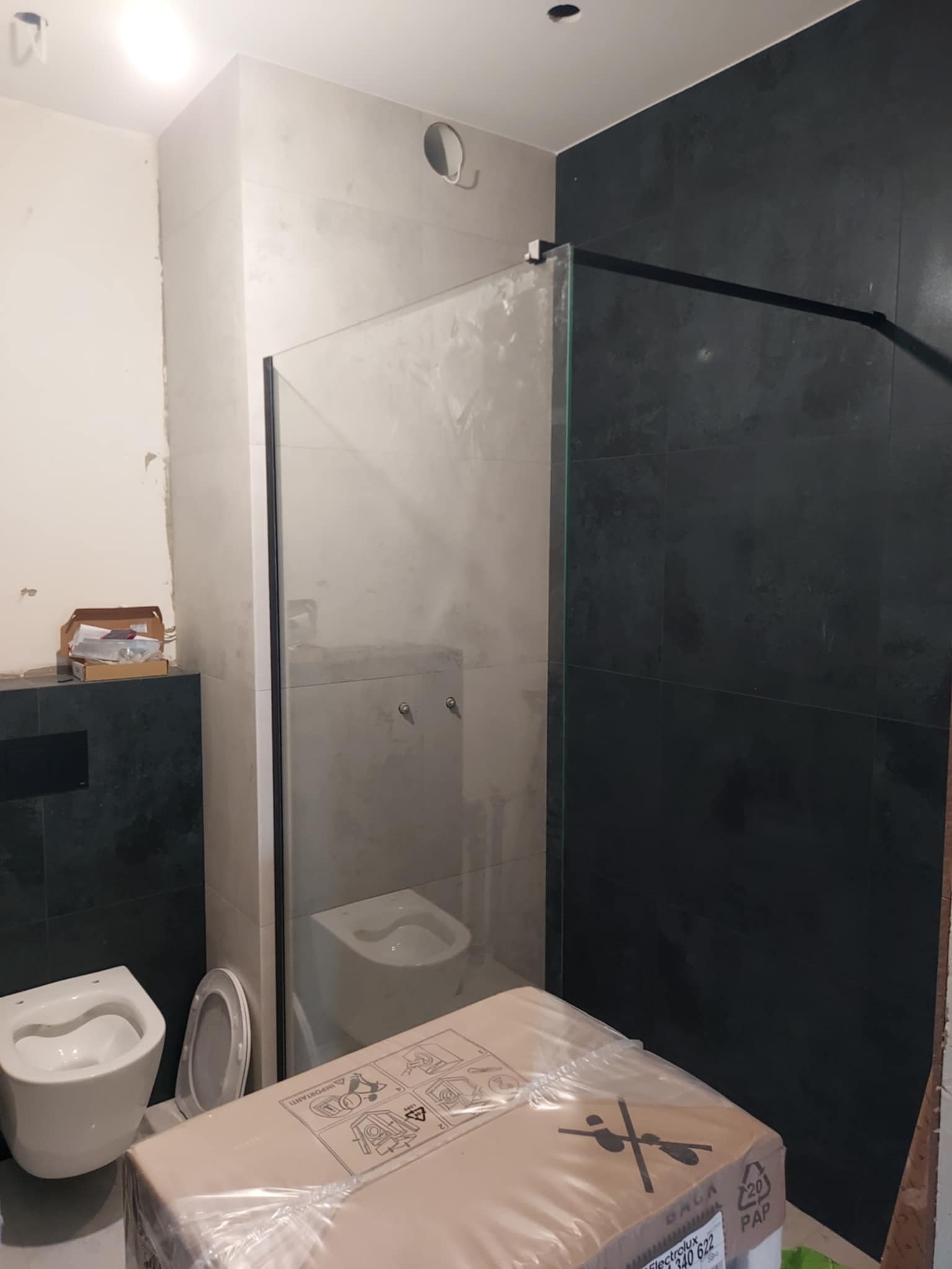 Łazienka w trakcie wykańczania: szklana kabina prysznicowa z czarną ramą, częściowo ułożone płytki w odcieniach szarości, toaleta w opakowaniu. Widoczne elementy instalacji.