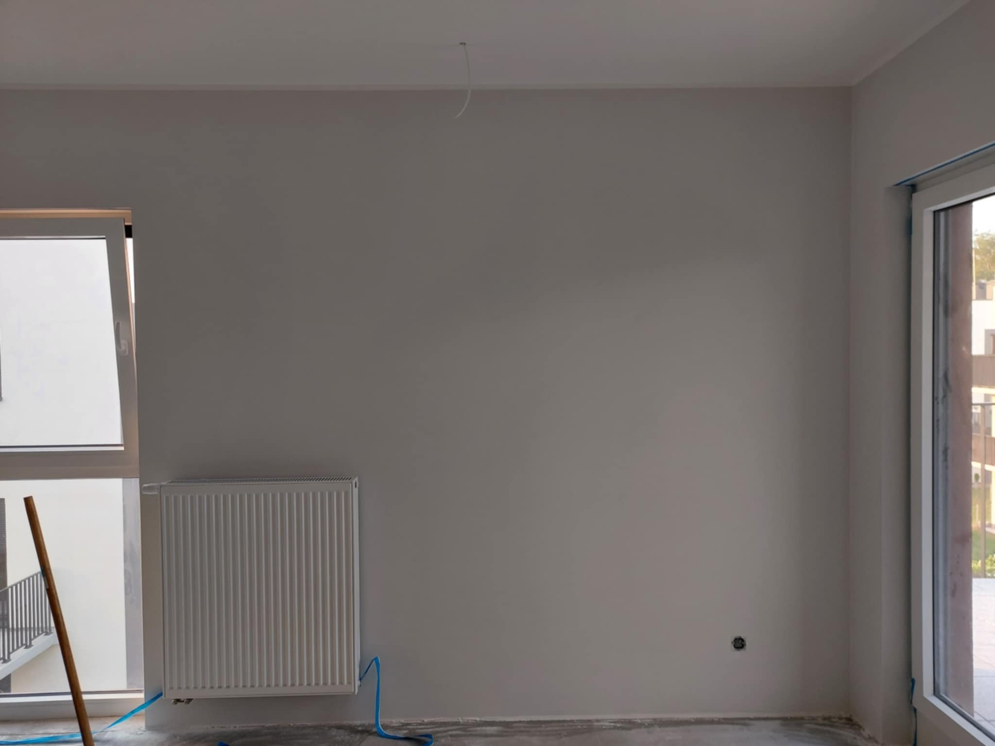 Wnętrze pokoju po szpachlowaniu ścian na gładko, z grzejnikiem i oknem. Widoczne ślady prac wykończeniowych na podłodze i wystający kabel ze ściany.