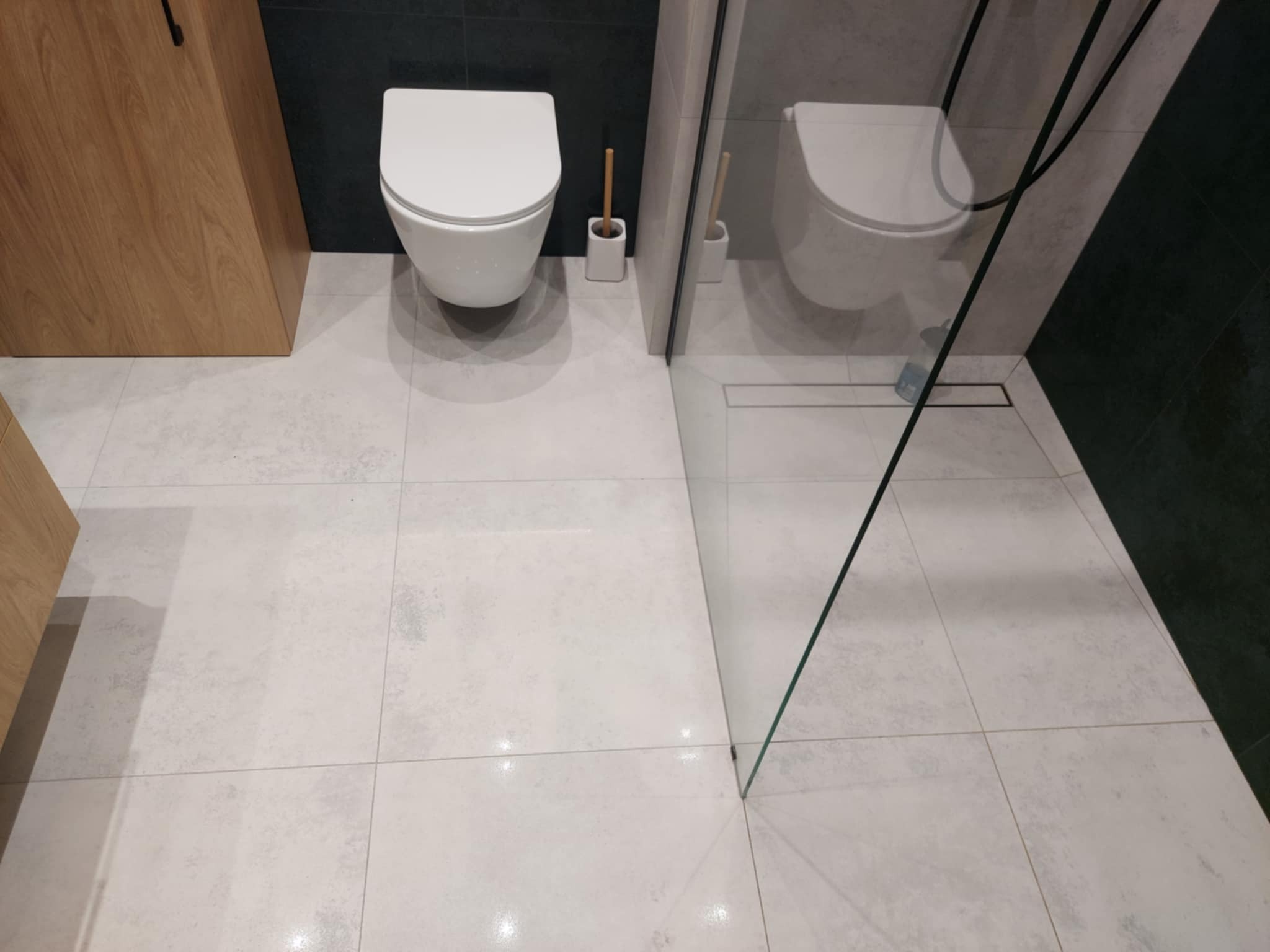 Nowoczesna łazienka z białymi płytkami, wiszącą toaletą i szklaną kabiną prysznicową. Minimalistyczny design z drewnianą szafką i czarnymi akcentami.