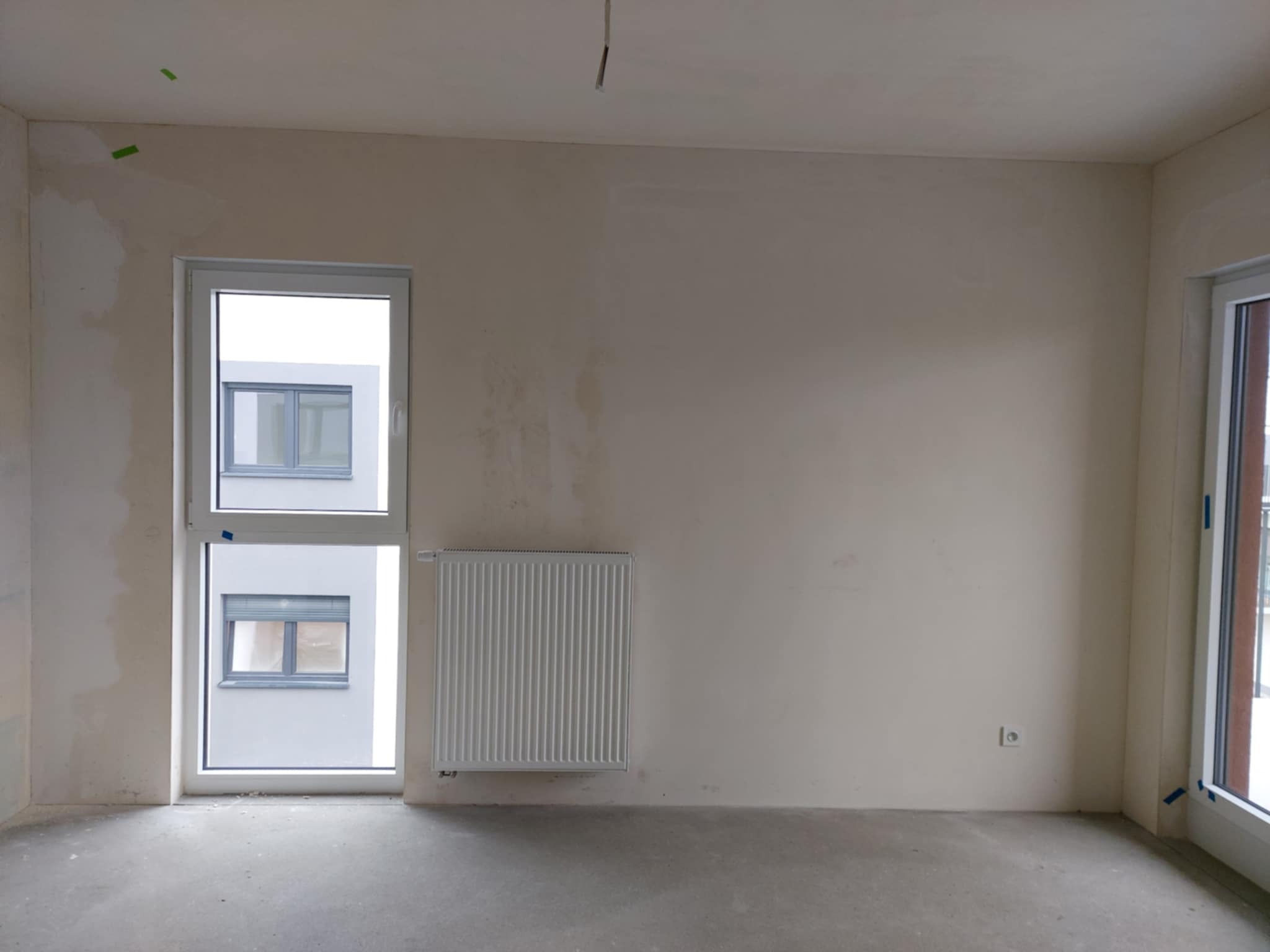 Pokój w stanie deweloperskim z grzejnikiem, surowymi ścianami z widocznymi śladami szpachlowania i oknami. Podłoga betonowa. Widoczne taśmy malarskie.