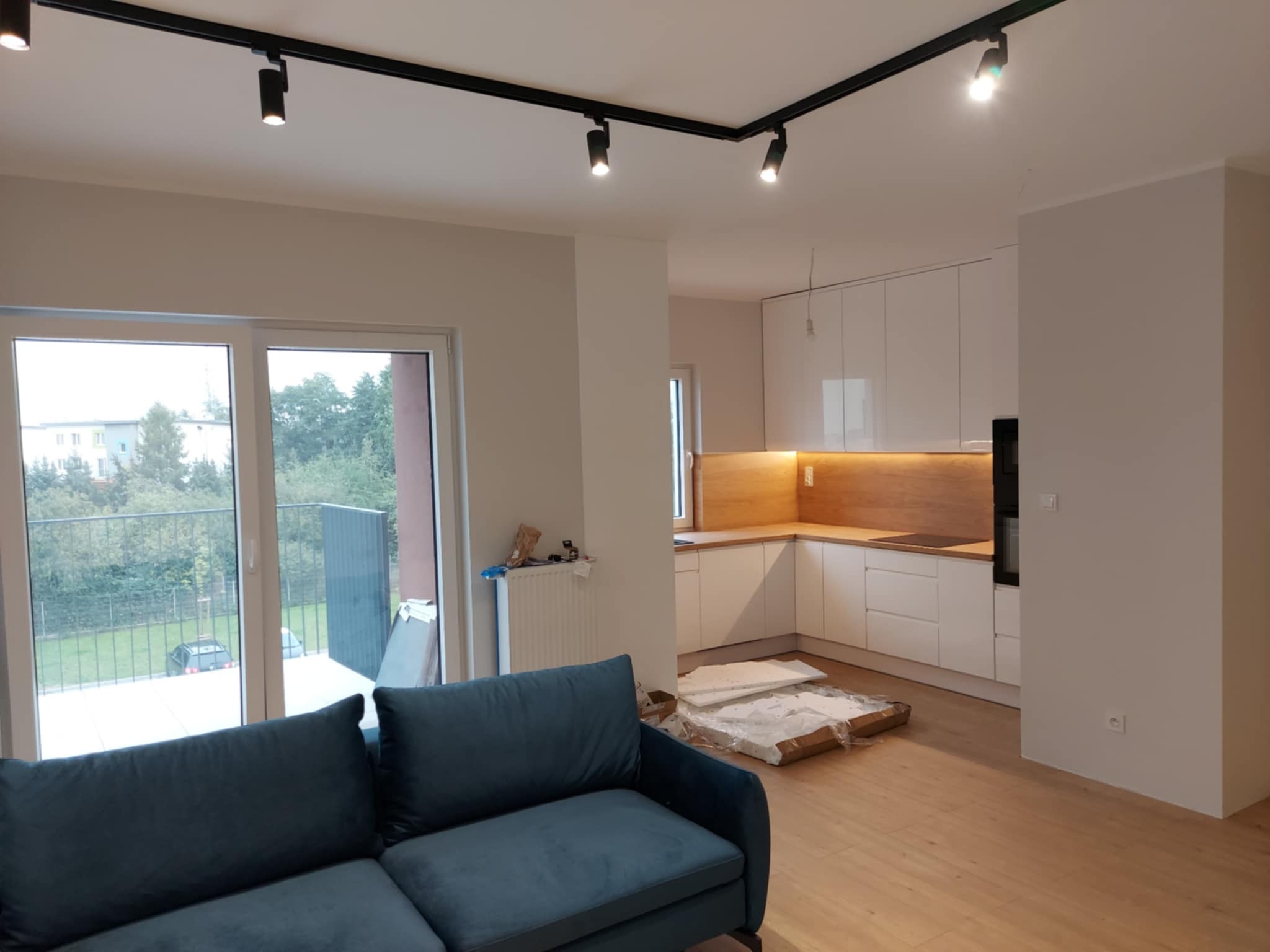 Nowoczesne wnętrze po remoncie: biała kuchnia z drewnianym blatem, oświetlenie szynowe, parkiet i niebieska sofa. Widok na balkon i zieleń za oknem.