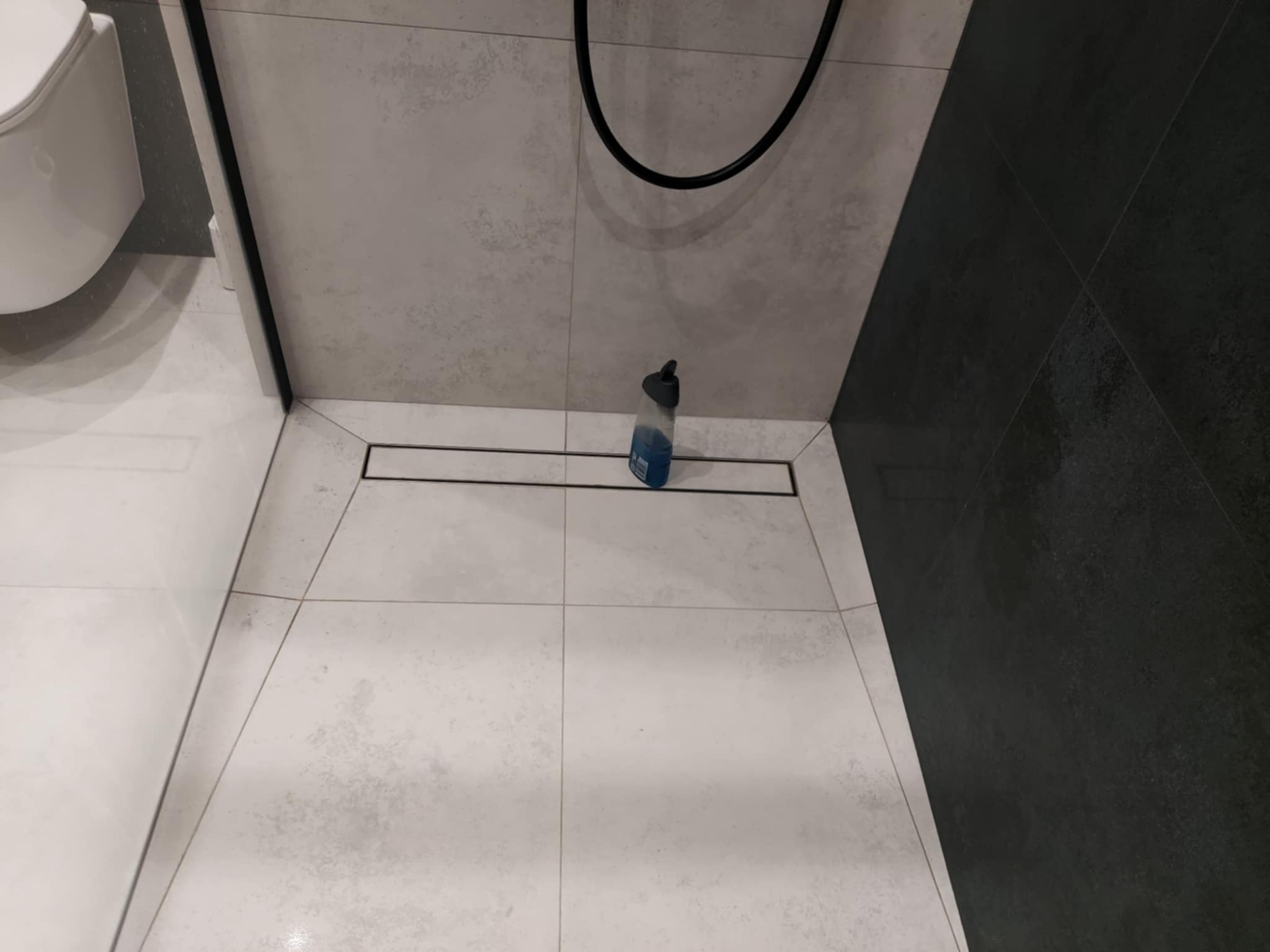 Nowoczesna kabina prysznicowa z odpływem liniowym, wykończona płytkami w odcieniach szarości. Widoczna butelka z płynem do czyszczenia.