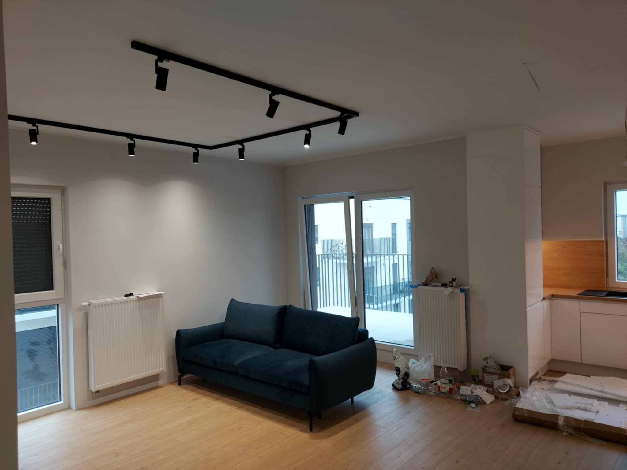 Jasne wnętrze z drewnianą podłogą, ciemną sofą i nowoczesnym oświetleniem szynowym. Widoczne okna balkonowe i grzejniki. Częściowo widoczna zabudowa kuchenna.
