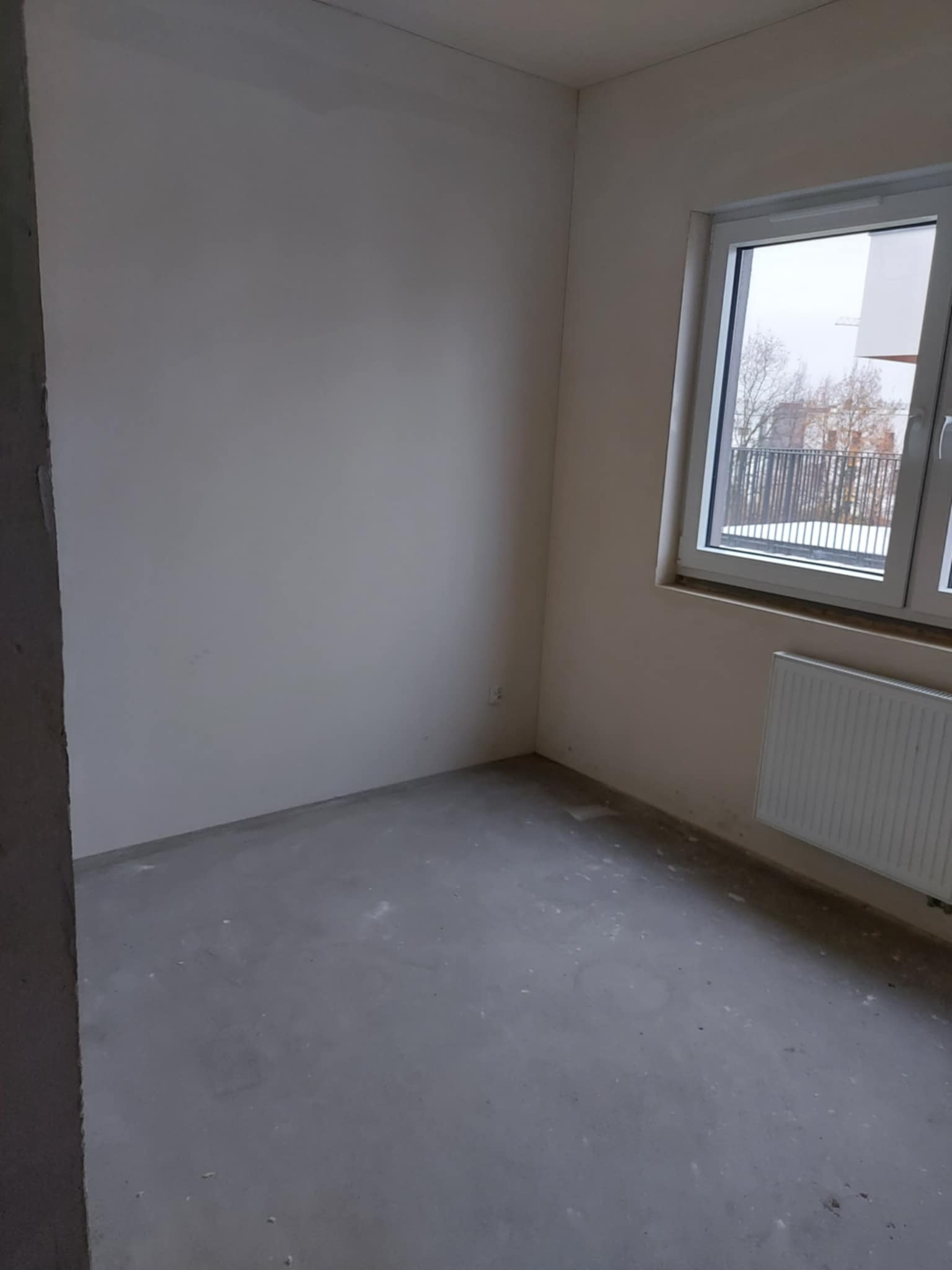 Pusty pokój z oknem i grzejnikiem, ściany przygotowane do malowania, surowy beton na podłodze. Widok na zewnątrz przez okno.