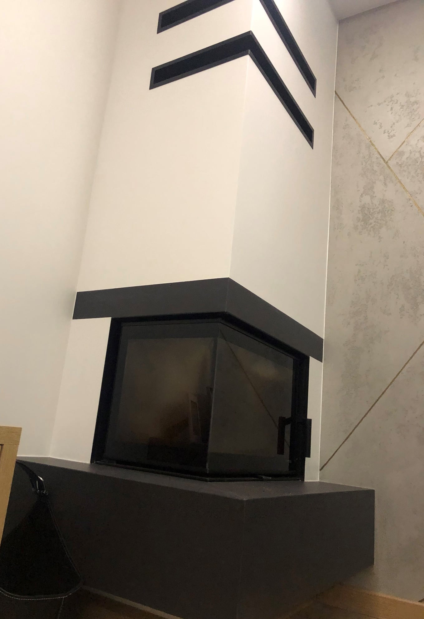 Biały kominek z czarnymi akcentami i geometrycznymi wnękami, obok ściana z szarymi płytkami i złotymi liniami. Nowoczesny design, minimalistyczny styl.