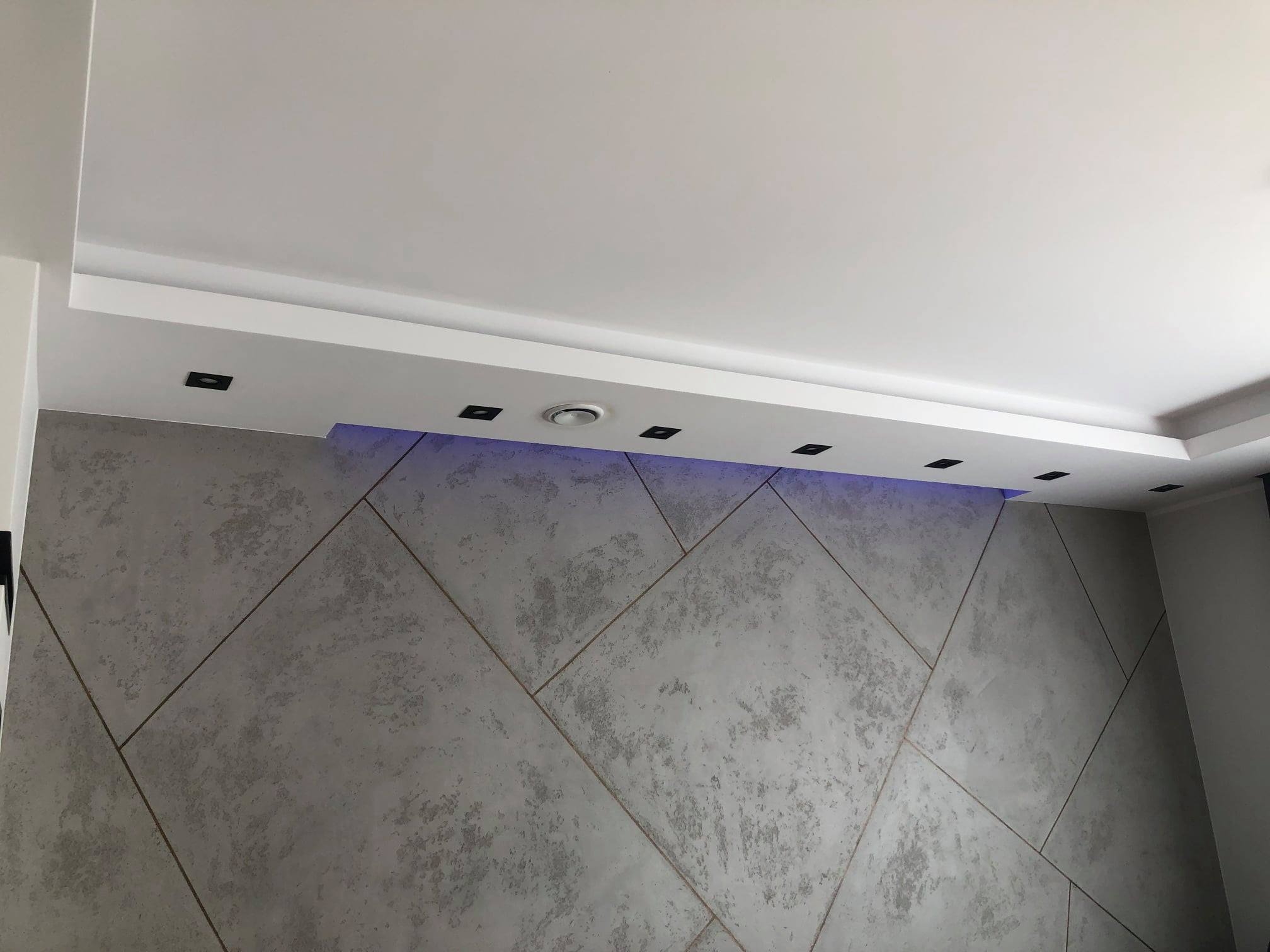 Dekoracyjna ściana z szarego betonu architektonicznego, podzielona geometrycznymi liniami, podświetlona subtelnym, niebieskim światłem LED w suficie podwieszanym.