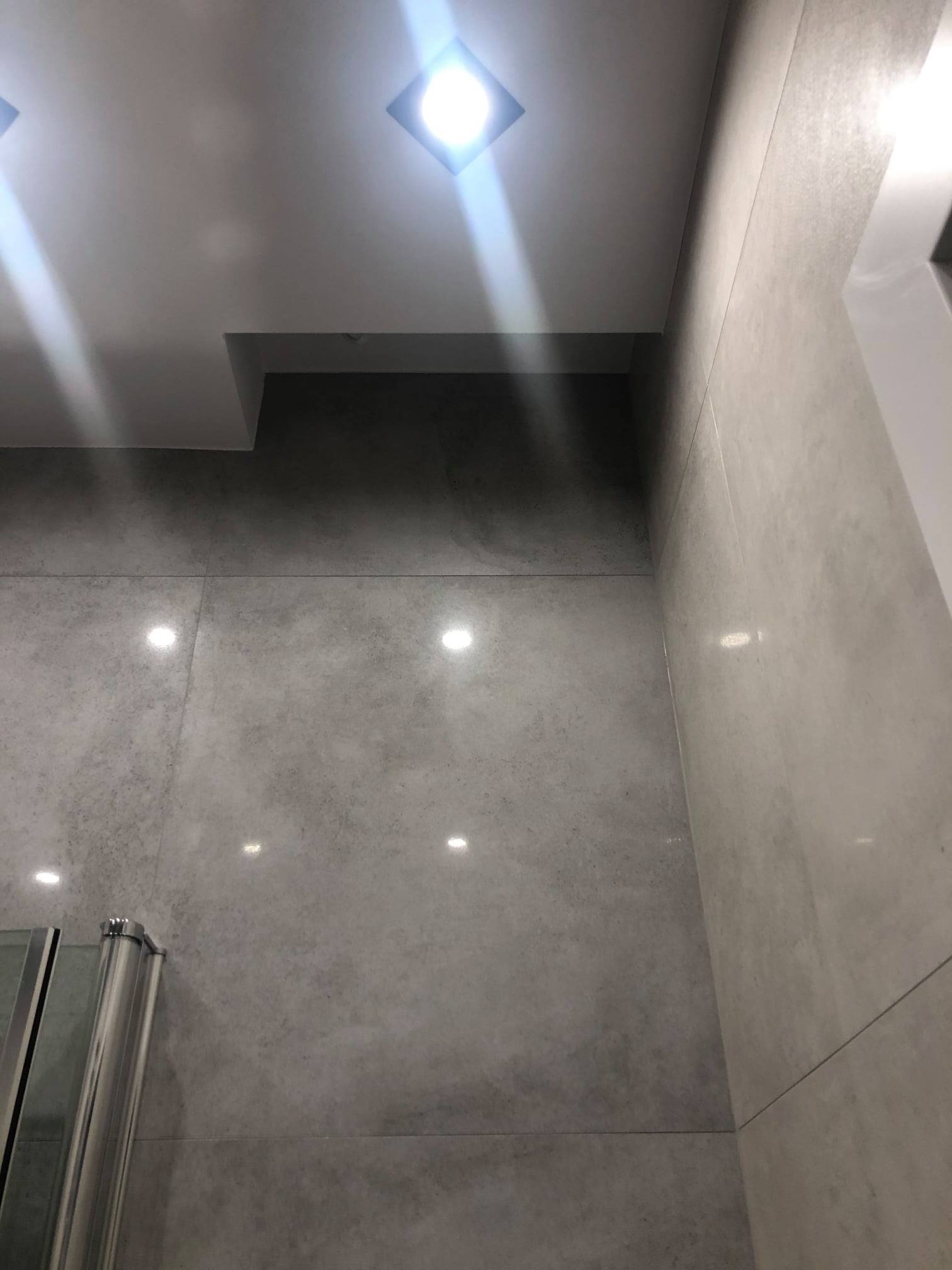 Szara, minimalistyczna łazienka z dużymi płytkami i punktowym oświetleniem sufitowym. Fragment kabiny prysznicowej widoczny w lewym dolnym rogu.