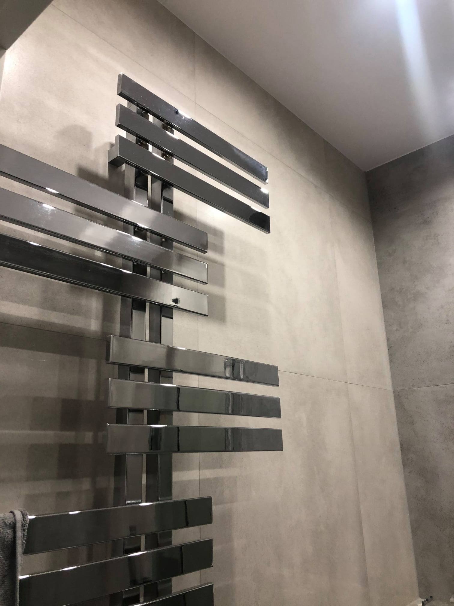 Nowoczesny, chromowany grzejnik łazienkowy o geometrycznym designie na tle płytek w odcieniach szarości. Minimalistyczne wykończenie wnętrza.