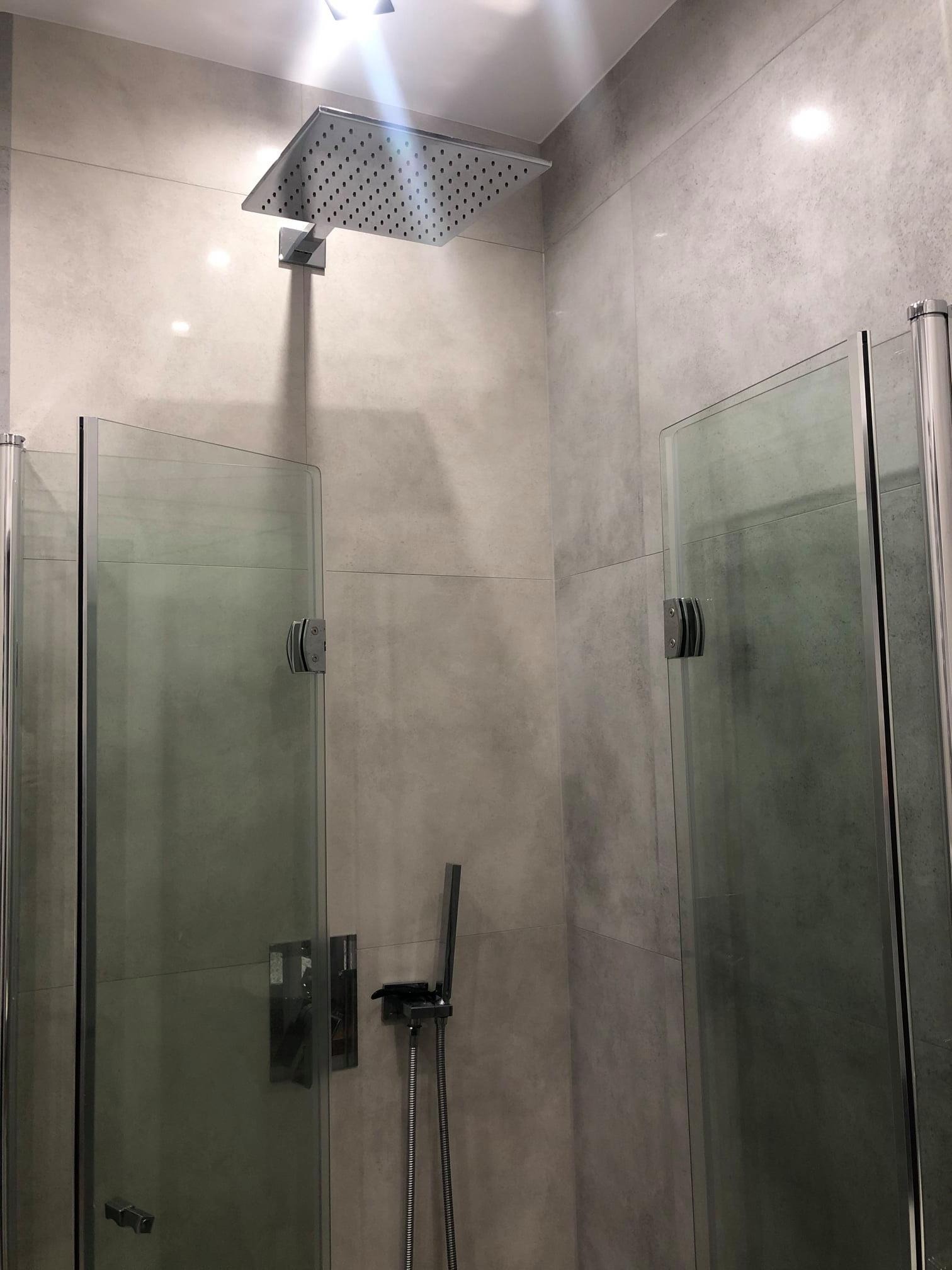 Nowoczesna kabina prysznicowa z deszczownicą i szklanymi drzwiami, wykończona szarą, matową płytką. Minimalistyczny design, widoczne detale armatury.