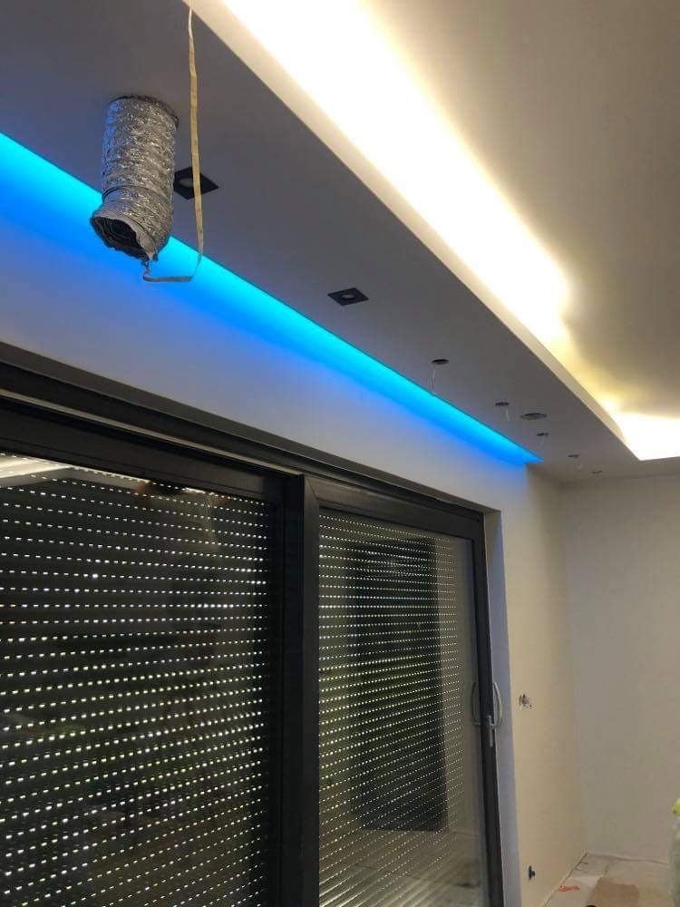 Nowoczesny sufit podwieszany z oświetleniem LED: niebieska listwa i ciepłe światło. Widoczne rolety okienne i fragment instalacji wentylacyjnej.