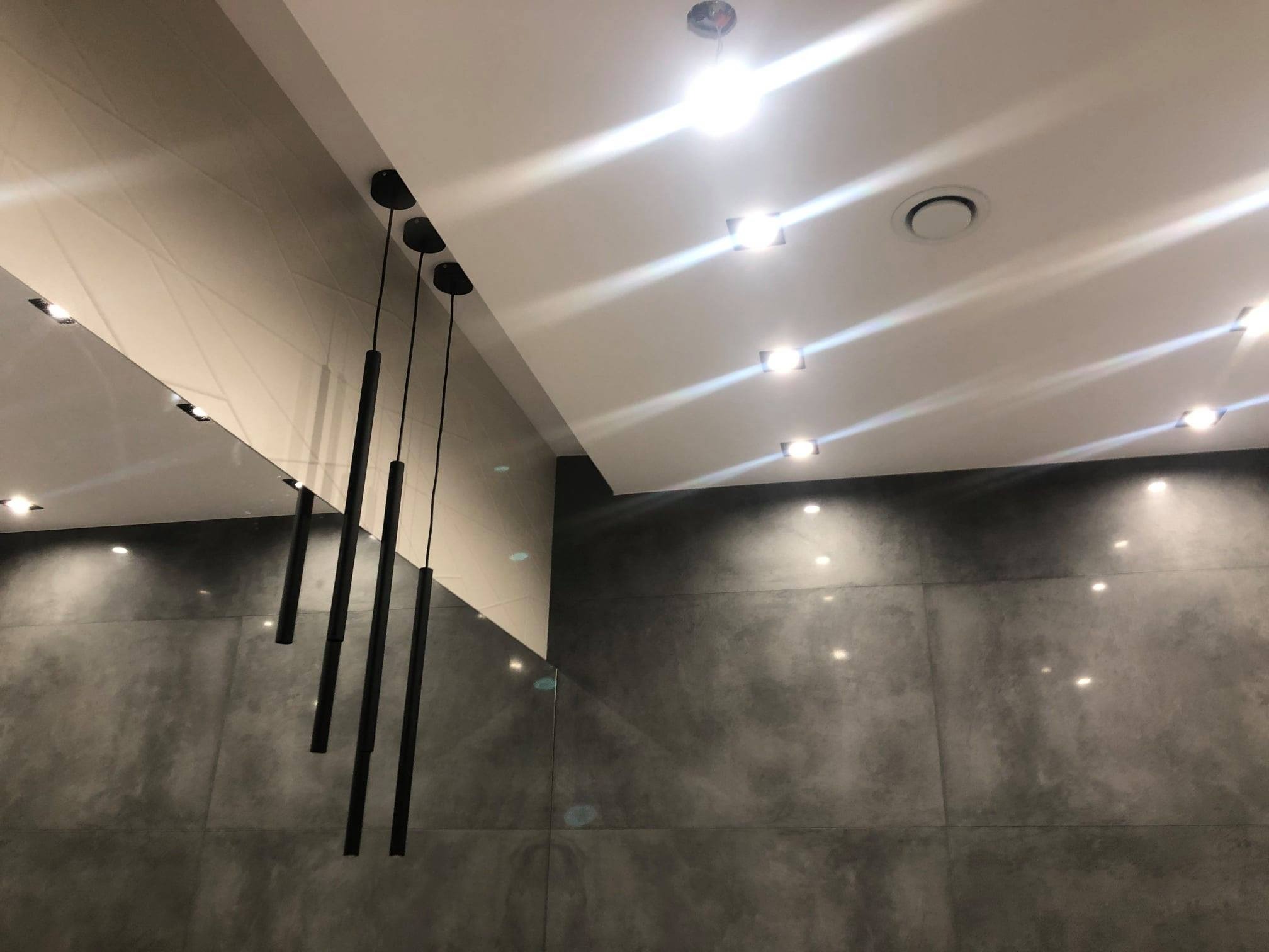 Nowoczesne wnętrze z szarą, betonową ścianą, oświetleniem punktowym i wiszącymi lampami tubami. Minimalistyczny design, sufit z geometrycznym wzorem.