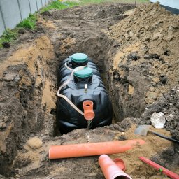 Firma Wielobranżowa Szymon Swacha - Czarny zbiornik z zielonymi pokrywami w wykopie, rury kanalizacyjne obok. Widok z góry na etap instalacji, narzędzia i ziemia w tle. Prace ziemne.