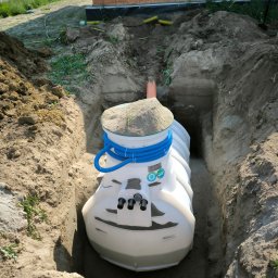 Firma Wielobranżowa Szymon Swacha - Montaż przydomowej oczyszczalni ścieków: biały zbiornik w wykopie, niebieskie rury, widoczny fragment budynku w tle. Etap instalacji.