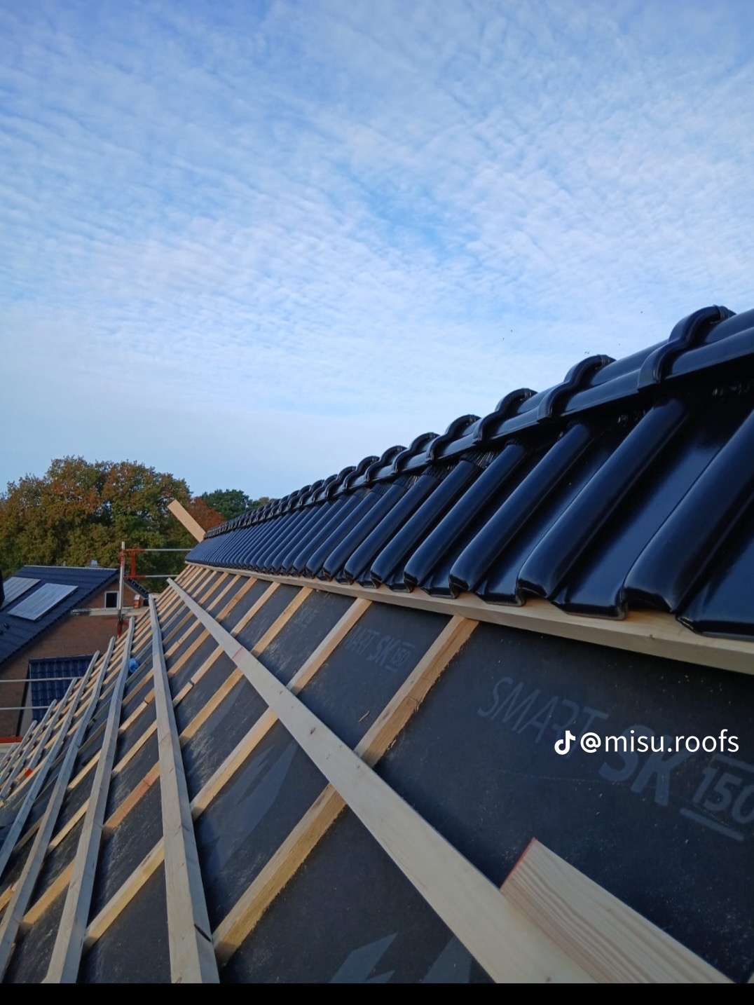 Częściowy widok dachu w trakcie krycia czarną dachówką, widoczne łaty i membrana dachowa. W tle błękitne niebo z chmurami i fragmenty innych budynków.