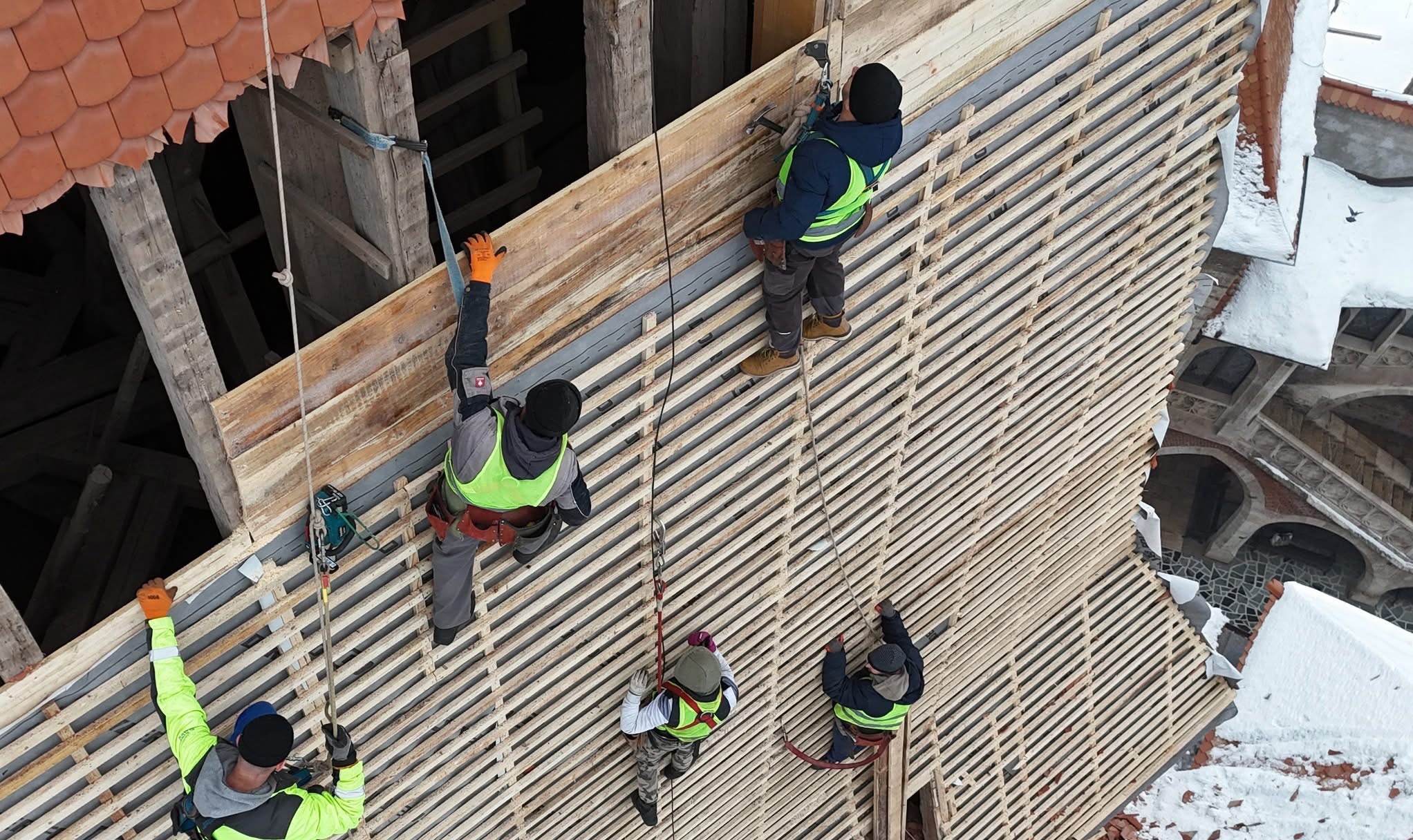 Ekipa monterów zabezpieczona linami, pracuje na drewnianej konstrukcji dachu z częściowo ułożoną dachówką. Widok z góry na zewnątrz budynku w zimowej scenerii.