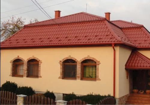 Dom z nowym, czerwonym dachem z dachówki ceramicznej, widoczne rynny i obróbki blacharskie. Elewacja w odcieniach beżu z ceglanymi detalami okiennymi.