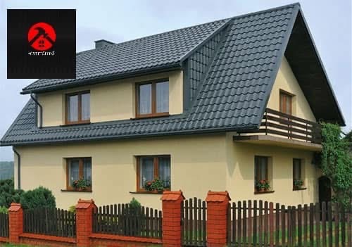 Dwupiętrowy dom z ciemnoszarym dachem z dachówki, brązowymi oknami i balkonem. Jasna elewacja, ogrodzenie z ceglanymi słupkami i drewnianymi przęsłami.