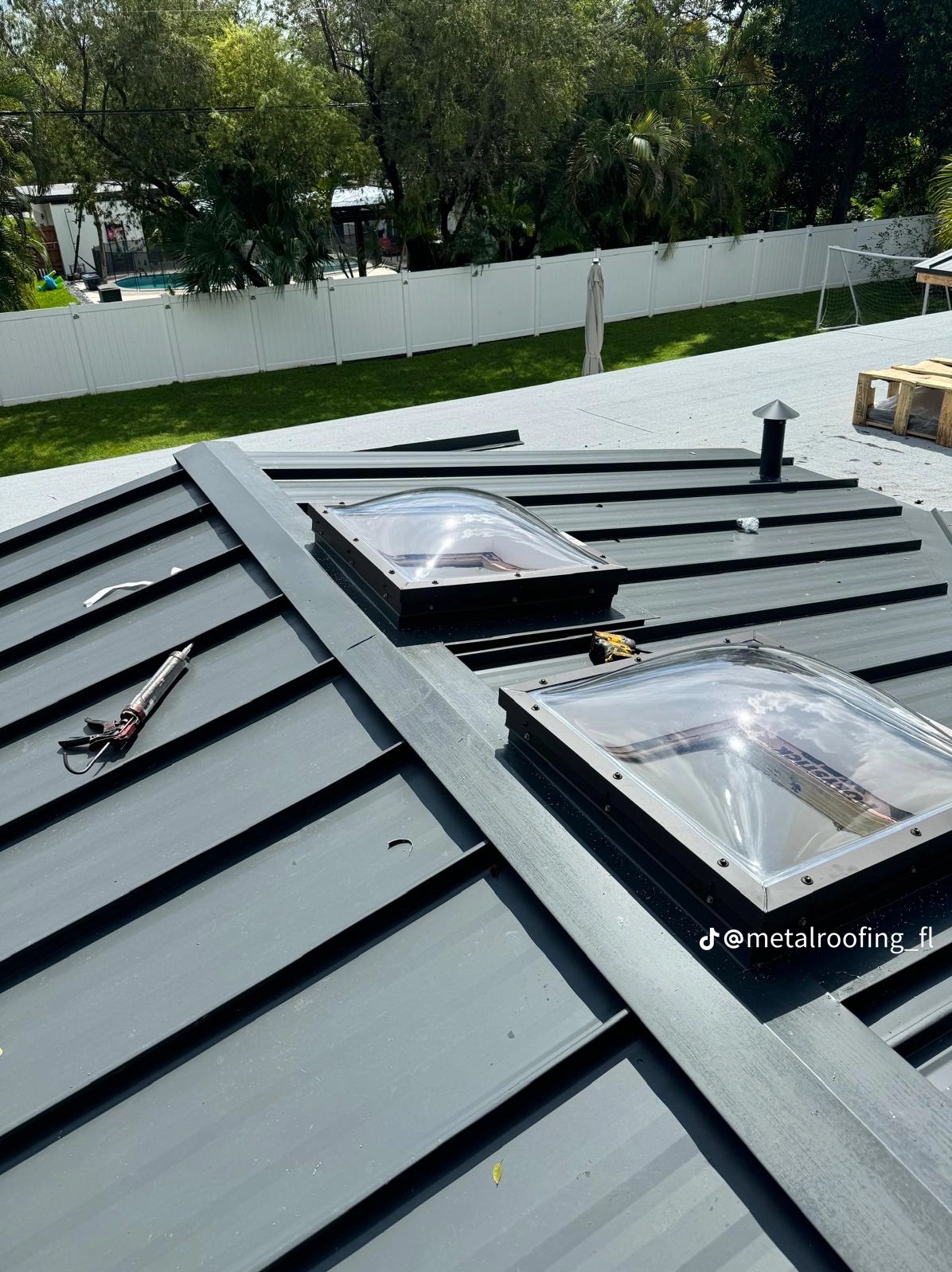 Szary dach z blachy trapezowej z dwoma świetlikami, narzędzia dekarskie w kadrze. W tle białe ogrodzenie i zieleń. Ujęcie z góry, w słoneczny dzień.