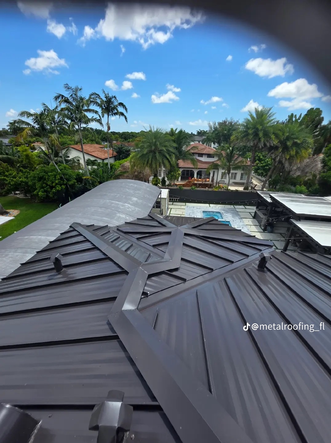 Nowoczesny, ciemny dach metalowy z widokiem na luksusową rezydencję z basenem i palmami w słoneczny dzień. Charakterystyczne łączenia blacharskie.