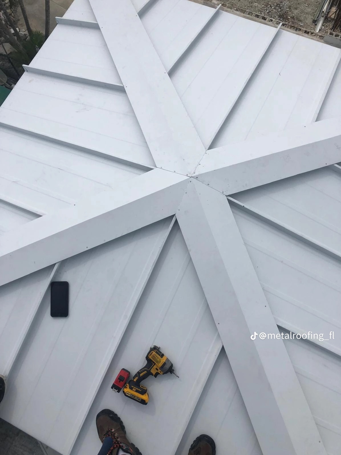 Biały dach z blachy trapezowej z widocznym narzędziem (wkrętarka) i telefonem. Ujęcie z góry, ukazujące symetryczny wzór łączeń blachy.