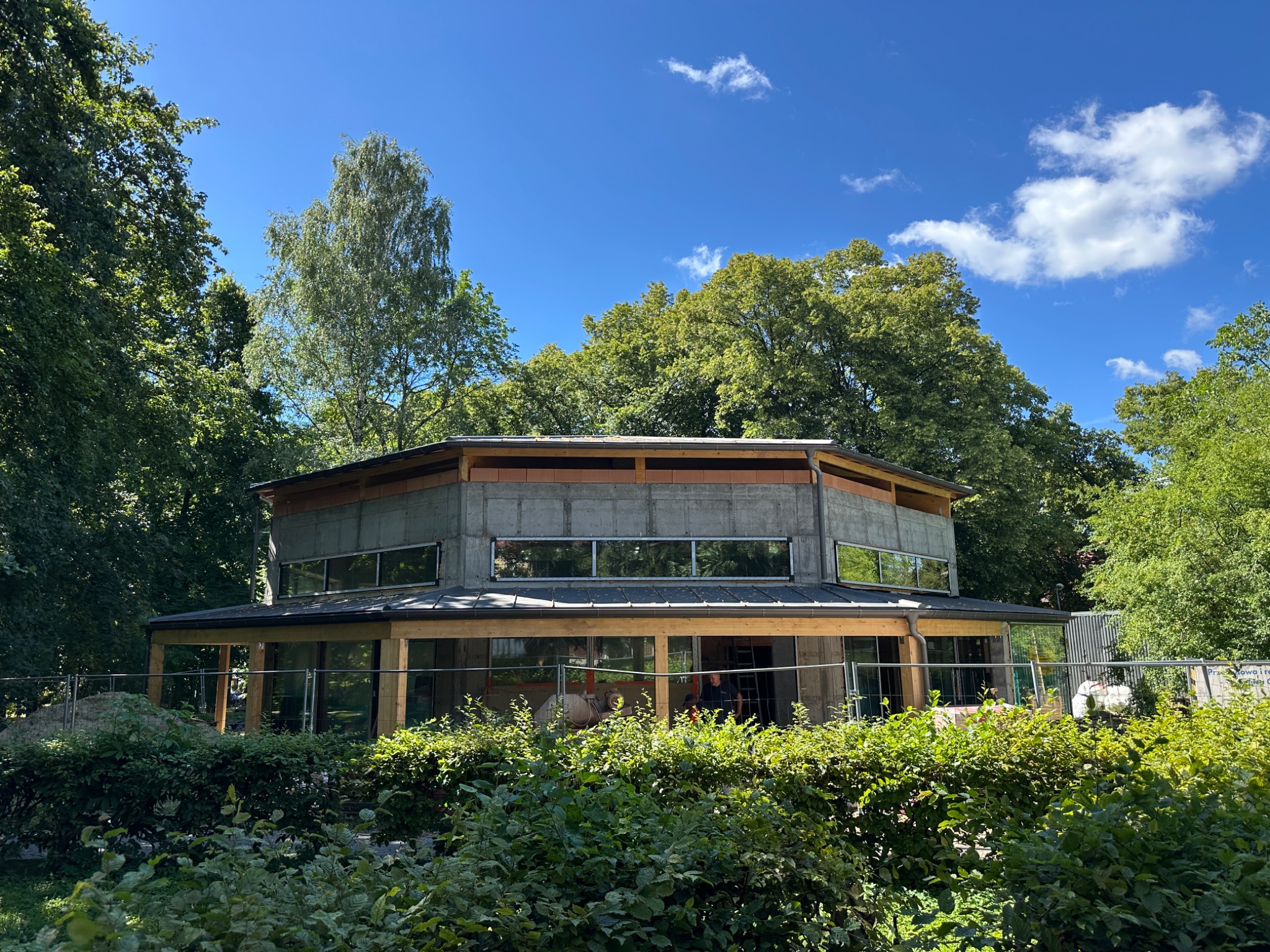 Nietypowy, okrągły budynek w trakcie budowy, otoczony zielenią. Widoczne surowe ściany, drewniana konstrukcja dachu i okna. Częściowo zasłonięty przez krzewy.