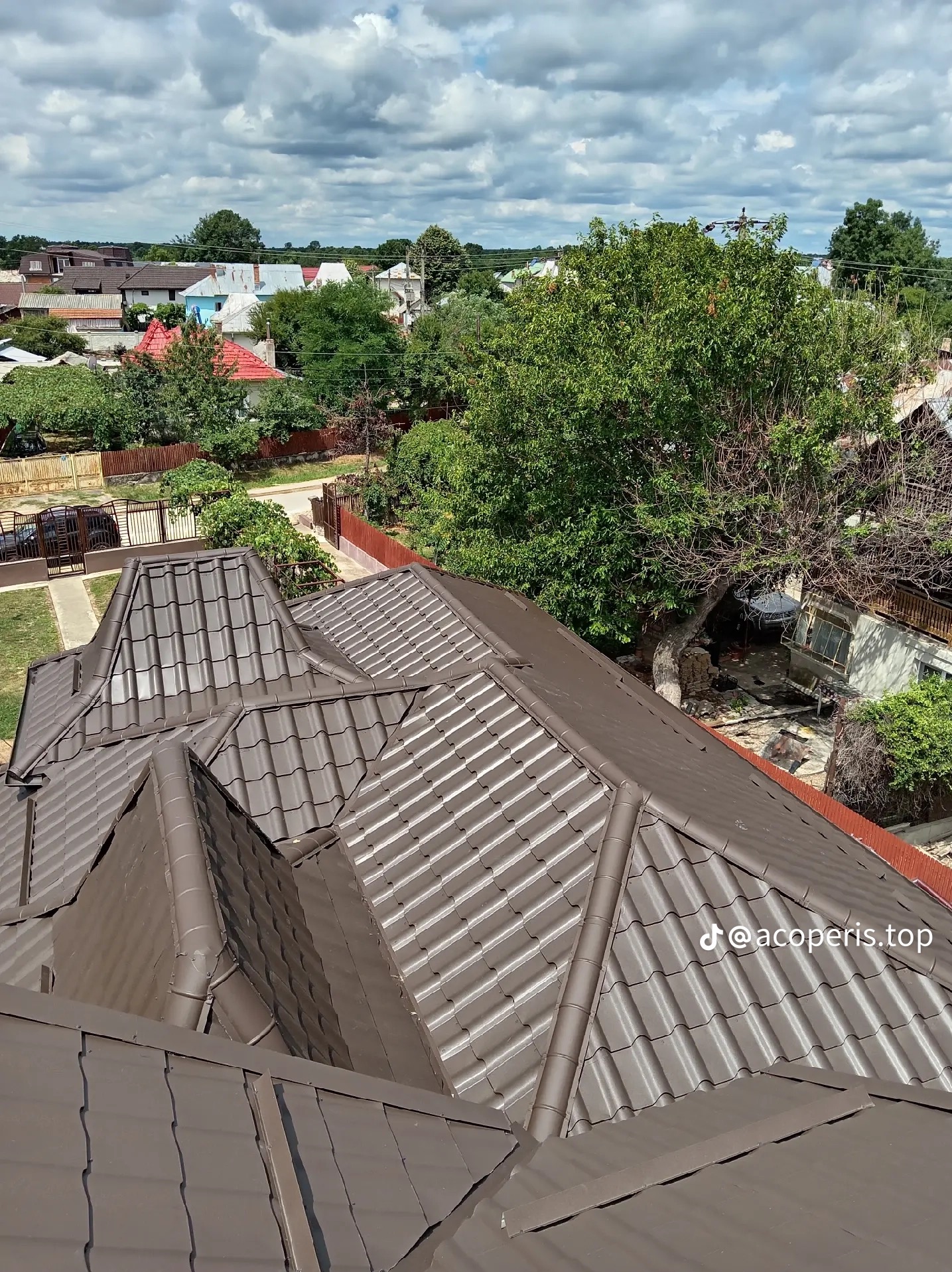 Nowy, brązowy dach z dachówki ceramicznej, widok z góry na tle zabudowy jednorodzinnej i zieleni. Widoczny fragment rynny i obróbki blacharskiej. Dach po renowacji.