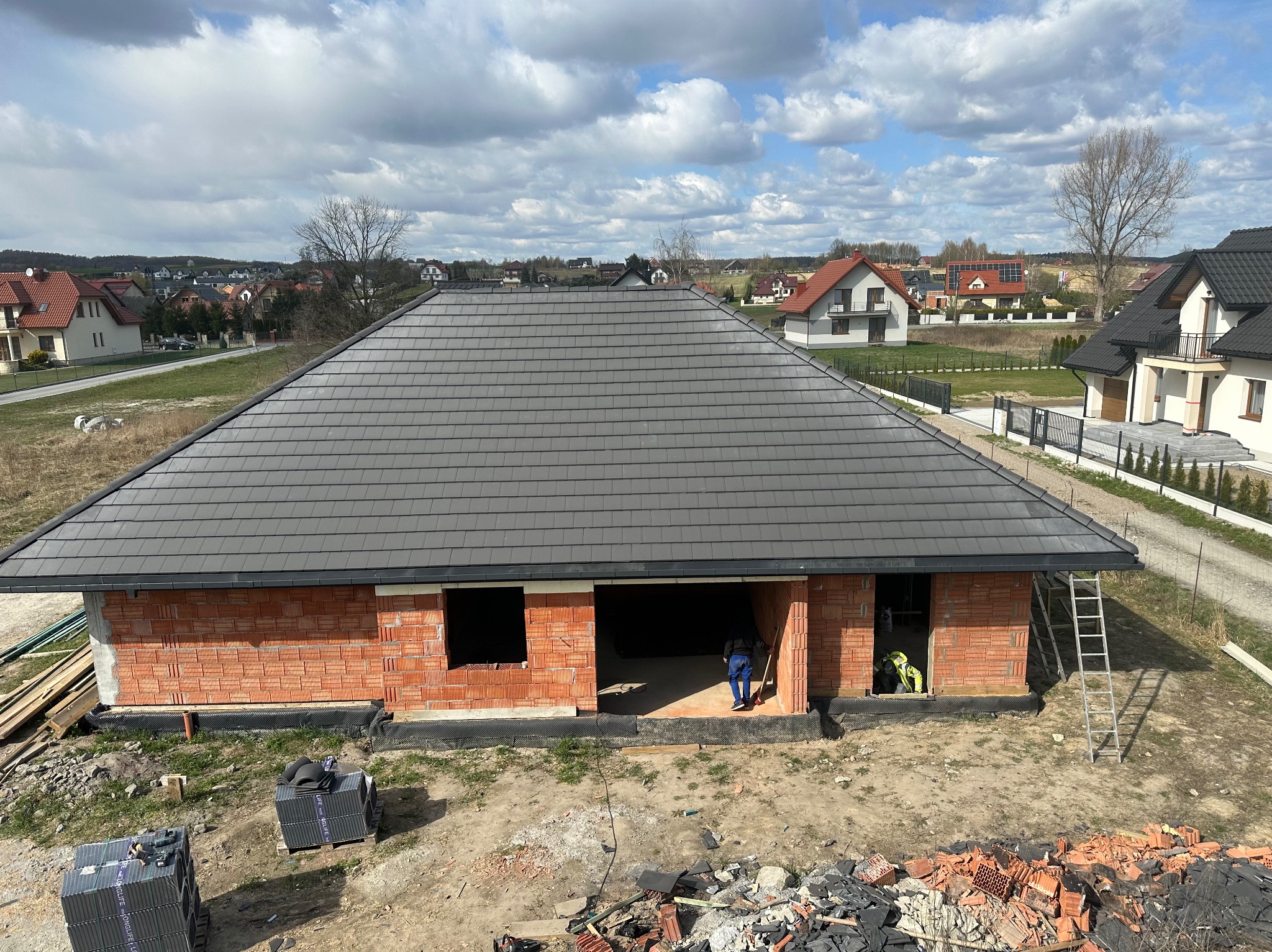 Nowy, szary dach z dachówki ceramicznej na surowym budynku z czerwonej cegły. W tle zabudowa jednorodzinna i pochmurne niebo. Widoczne palety z dachówką i drabina.