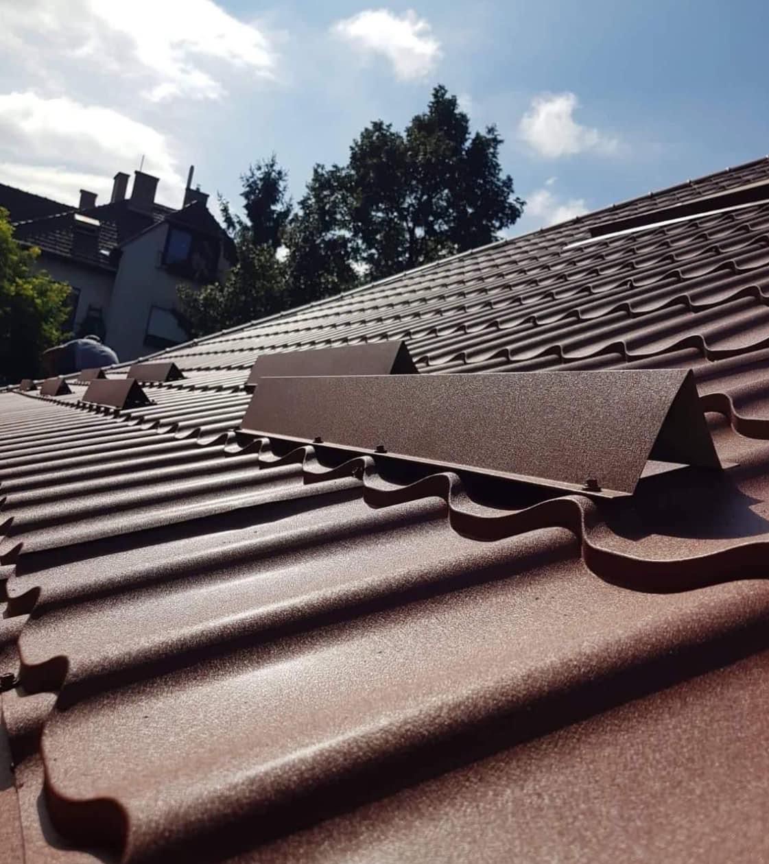 Brązowy dach z blachodachówki z zamontowanymi elementami wentylacyjnymi, widok pod kątem, błękitne niebo w tle. Detal nowego pokrycia dachowego.