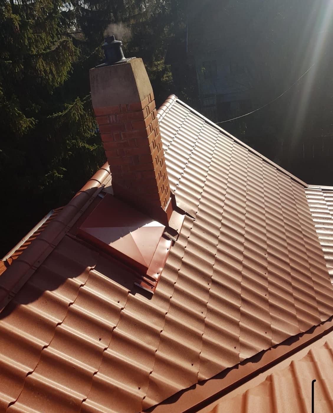 Ceglasty dach z kominem w Katowicach, widok z góry. Nowoczesna dachówka ceramiczna, obróbki blacharskie wokół komina. Słoneczny dzień, drzewa w tle.